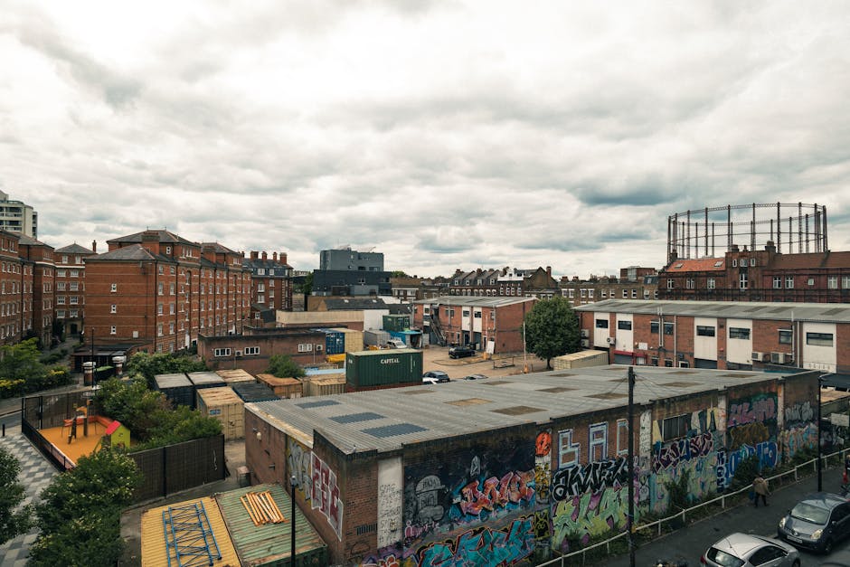 A daytime urban scene showing a mix of residential and commercial buildings under a cloudy sky. In the foreground, there is a building with a flat, corrugated metal roof decorated with colorful graffiti artwork along its sides. Surrounding this building are small trees, a fenced playground with a slide and play area, and parked cars along the street. Behind, several multi-story red-brick residential blocks and industrial-style structures are visible, including a large, open framework of a circular structure on top of one building. Additional warehouses, shipping containers, and an assortment of smaller structures are scattered throughout the scene. The environment is typical of a busy cityscape with a combination of utilitarian and decorative elements, illustrating a context where private waste management might be carried out in addition to municipal services, aligning with typical rubbish removal and on-site clearance activities in dense urban areas.