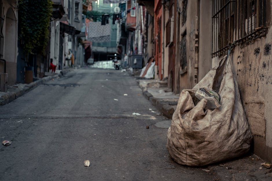 A large, weathered beige plastic rubbish bag filled with waste rests on the uneven, dark grey asphalt pavement of a narrow urban street. The bag appears slightly dirty, with creases and minor tears visible on its surface, and a partially visible transparent plastic container protrudes from its top. Surrounding the bag, small pieces of litter such as crumpled paper and wrappers are scattered across the ground. On the right side, there is a brick building with a rusted metal security grille over a window, and the wall is covered in faded graffiti and grime. The street extends uphill, lined with tightly packed residential buildings constructed from various materials including brick and concrete, some with peeling paint or exposed surfaces. In the distance, laundry hangs on lines stretched between buildings, and a few pedestrians and parked bicycles can be discerned, contributing to the bustling atmosphere typical of an urban environment where private waste collection services, like those offered by Waste Collection Hackney, often handle rubbish removal on narrow streets that pose access challenges for standard bin lorries. The scene is lit with diffuse, natural daylight, emphasizing the gritty, lived-in character of the street.