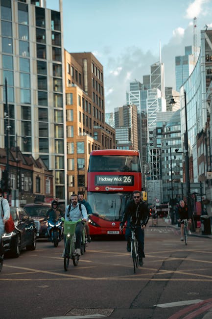 A busy urban street scene in Hackney featuring a red double-decker bus displaying the route number 26 and the destination 'Hackney Wick' in illuminated digital signage at the front. The bus is centrally positioned, with several pedestrians and cyclists in the foreground, including a man wearing sunglasses and a dark jacket riding a bicycle, and others walking or cycling nearby. The surrounding environment includes modern glass-fronted office buildings and older brick structures on either side of the street, with the sky showing a partly cloudy blue tone. Vehicles are parked along the sides, and the scene captures the vibrancy of city life with a mix of transportation modes and architectural styles. Waste Collection Hackney provides private rubbish removal, supporting alternative waste handling within this busy commercial and residential area, with visible infrastructure indicating active urban management.