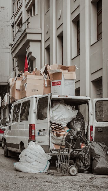 A white commercial van with its rear doors open, parked along a city street adjacent to a multi-story building with beige masonry. The van's interior is filled with large black and white garbage bags, cardboard boxes, and wooden pallets, indicating the collection of bulky waste for disposal. Several cardboard boxes, some open and damaged, are stacked on top of the van, with visible labels and varying degrees of wear and tear, suggesting recent loading. In front of the vehicle, additional black and white garbage bags and discarded materials are on the pavement, alongside a small metal trolley. The scene captures an urban environment typical of on-site or private waste collection, where waste is temporarily stored or prepared for removal. The surrounding area features the building’s facade with rectangular windows and decorative architectural details. The lighting appears natural, with overcast sky conditions creating diffuse illumination, highlighting the textures and materials of the discarded rubbish and the vehicle. Waste Collection Hackney's service is implied by this scene of independent rubbish clearance involving bulky waste handling near commercial or residential premises.