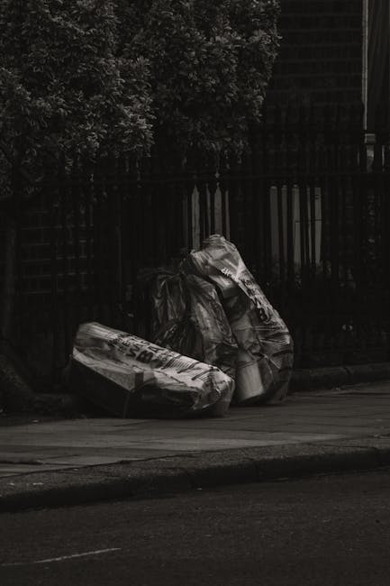 Several large black plastic refuse bags, some slightly crumpled and others more tightly sealed, are placed on the pavement near a black metal fence. The bags appear to be filled with household waste or discarded materials. Behind the fence, dense greenery is visible, with leafy branches extending overhead. The scene is set in an outdoor urban environment, with the bags positioned at the edge of a sidewalk adjacent to a road or driveway. The lighting suggests a cloudy or shaded day, with minimal natural light illuminating the scene. The bags' surfaces have a glossy finish, and they are stacked loosely with some leaning against the fence or each other. This setup reflects an instance of private or independent rubbish disposal, possibly arranged through waste collection services such as Waste Collection Hackney, to handle general waste or bulky refuse outside designated municipal collection areas.