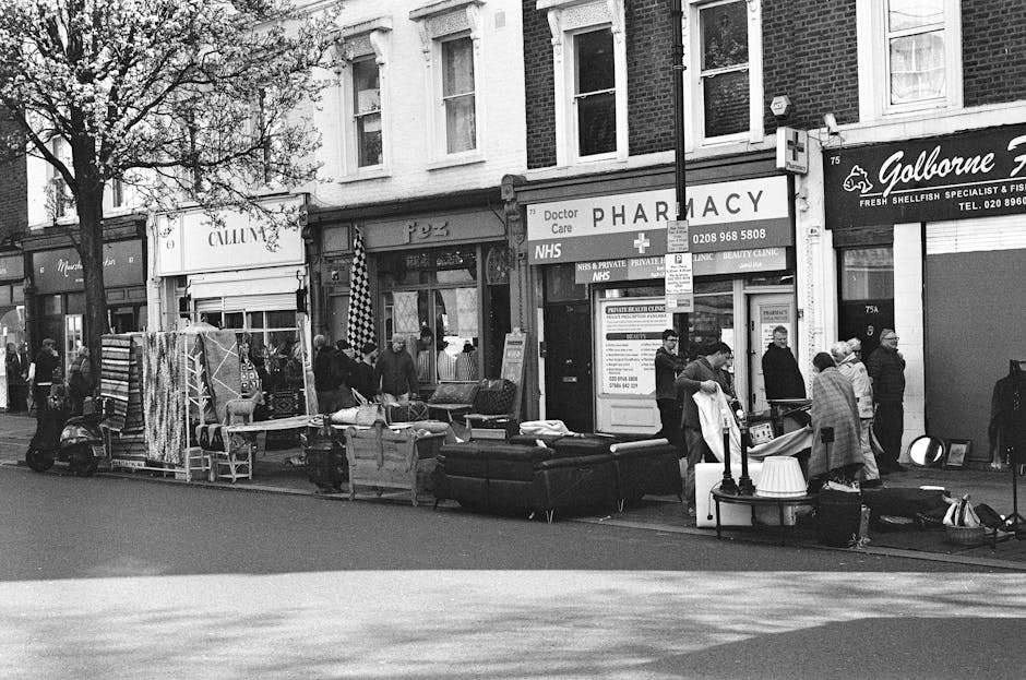 A street scene in front of a row of shops and a pharmacy, with people gathered around a variety of discarded furniture and household items placed along the pavement. Visible objects include a dark-colored sofa, a small table with a lamp, a stack of fabric or rugs, and other miscellaneous household goods, arranged as part of a clearance or disposal of unwanted items. The furniture appears to be in good condition, with textured upholstery on the sofa and smooth surfaces on the tables and lamps. Several individuals are engaged in examining or handling the items, while others stand nearby, possibly waiting or observing. The surroundings feature a brick building with shopfronts, including a pharmacy with signs and window displays, and a vertical pole with street signs. The overall scene is shaded by a large tree, and the natural daylight suggests an outdoor environment utilising direct sunlight, typical for private or third-party rubbish removal activities on urban streets, emphasizing a context where independent collection or on-site clearance of household waste and bulky items is taking place.