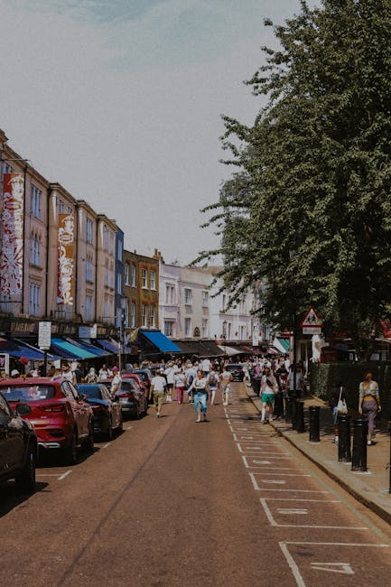 A busy street scene at Columbia Road in Hackney, showing a row of multicolored, closely packed terraced buildings with retail shops and market stalls along the pavement. The market stalls are covered with dark-colored awnings, and numerous pedestrians are walking along the street, some carrying shopping bags or engaged in conversation. On the left side, a line of parked cars is visible, with a narrower lane designated for vehicles, marked with white lines and numbers. The right side features tall, leafy trees providing shade, with park bollards separating the walkway from the street. The scene captures an outdoor market environment with a lively atmosphere, and the visible rubbish removal involves accumulated waste and discarded items likely cleared by private waste collection services such as Waste Collection Hackney, supporting efficient on-site clearance and alternative waste handling solutions at a busy market location.