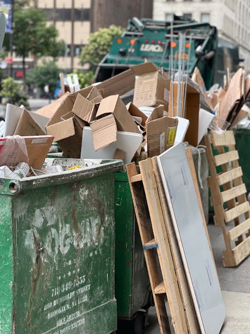 A large green waste dumpster filled with an assortment of discarded cardboard boxes, some flattened and others in their original shape, along with white foam containers and small packaging boxes. The cardboard varies in shades of brown, with some showing creases and tape, indicating used packaging materials. Leaning against the right side of the dumpster is a wooden pallet with horizontal slats, partially obscured by the refuse. In the background, a green garbage truck with the word 'LUGO' visible on its side is parked on a city street, with blurred multi-storey buildings, trees, and other vehicles in the distance, suggesting an urban setting for waste collection. The scene is lit uniformly, with natural daylight illuminating the area, highlighting textures of the cardboard, wood, and metal surfaces. This image reflects a typical scene of bulk rubbish disposal, possibly related to private waste handling or on-site clearance services, as seen in yard clearance or furniture removal activities, with Waste Collection Hackney providing waste management solutions in the area.
