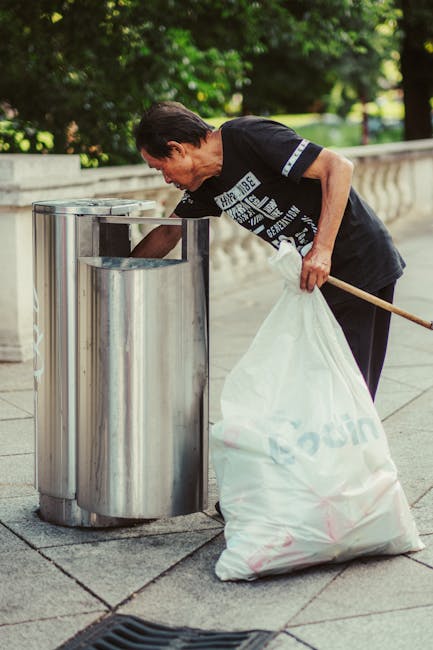 A middle-aged man with dark hair, dressed in a black T-shirt and dark trousers, is seen disposing of waste into a stainless steel public rubbish bin located on a paved footpath. He is leaning forward, holding a white plastic bag filled with garbage in his left hand, while using his right hand to place or retrieve rubbish from the top opening of the bin. The man is standing beside a waist-high, cylindrical, silver-colored waste container positioned on a concrete surface, with a curved, decorative stone balustrade visible in the background. Surrounding the scene are lush green trees and foliage, suggesting an outdoor park or pedestrian area during daylight. The environment appears clean and well-maintained, with natural lighting casting soft shadows. The image subtly reflects the concept of personal or independent waste disposal, aligning with practices of local waste collection or private rubbish removal services that may complement conventional public rubbish collection methods.