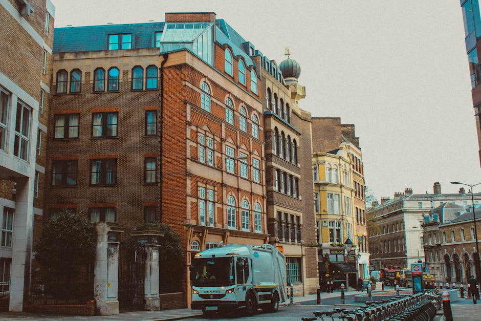 A street scene featuring a row of multi-story buildings with varied architectural styles, including a prominent red-brick structure with detailed stonework around its numerous windows and a decorative dormer window on the roof. The foreground displays a white waste collection truck with the branding of Waste Collection Hackney, parked adjacent to a curb, close to an entrance gated with ironwork and stone pillars. To the right, a yellow Victorian-style building with ornate bay windows and decorative elements adds historic character, while further down the street, more traditional and modern buildings line the pavement. The street is lined with bollards, and a series of empty bicycle racks is visible in the lower right corner. Overhead, the sky is overcast, casting diffuse light over the scene, which contextually aligns with alternative or private waste collection activities typical for urban environments where independent rubbish removal services might operate, such as those provided by Waste Collection Hackney in the E8 area of Hackney.