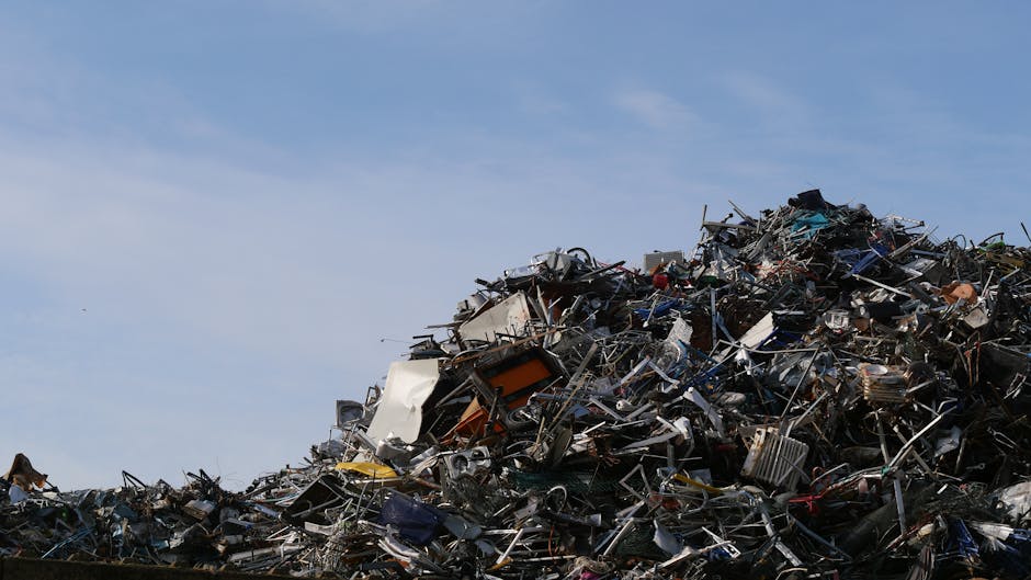 A large mound of mixed metal and electronic waste is piled high outdoors against a background of a clear blue sky. The debris includes various discarded appliances, metal frames, and electronic components, with some items exhibiting rusted or corroded surfaces. The materials appear weathered, with textures ranging from smooth painted surfaces to rough, jagged edges of bent or broken metal parts. The pile stretches diagonally from the lower left to the upper right of the image, occupying most of the foreground, while the sky provides a contrasting backdrop. The environment suggests an industrial or waste management site where waste materials are collected or sorted before further disposal or recycling. The scene is illuminated by natural daylight, highlighting the metallic sheen and textures of the clutter. As a professional waste management service, Waste Collection Hackney handles such waste, which may include non-compliant or illegally dumped items, underscoring the importance of proper rubbish removal practices to prevent unsightly and potentially hazardous accumulations.