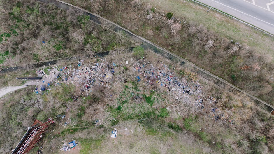 An aerial view of an outdoor area showing a large, irregularly shaped pile of mixed waste and rubbish, including plastic bottles, bags, paper, and other debris, scattered across a patch of bare ground with sparse vegetation. The surrounding environment features leafless trees and bushes, with a paved road running along the top edge of the image and a narrow concrete wall separating the waste area from the roadside. In the bottom left corner, there is a small section of red metal framework or equipment. The scene appears to depict a location where illegal dumping or unmanaged waste accumulation has occurred, highlighting the need for proper rubbish removal services to address illegal rubbish dumping in the area, an issue often linked to the activities managed by waste collection specialists such as Waste Collection Hackney.