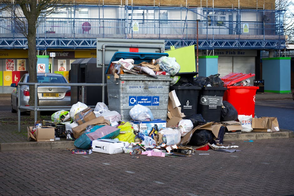 An overflowing waste bin with a blue lid and marked for mixed paper and cardboard is situated on a paved street, with various discarded items piled around its base. The debris includes cardboard boxes, plastic bags, crumpled paper, and packaging materials, some spilling onto the nearby curb. The waste appears to be a mixture of household rubbish, with both black and black-and-white plastic trash bags, as well as loose items, contributing to the cluttered scene. In the background, there is a metal railing, a parked silver car with a yellow license plate, and a building under construction or renovation, partially covered with scaffolding and blue safety netting. The environment suggests a typical urban area where bin overflow may occur, necessitating private waste collection services such as those provided by Waste Collection Hackney, especially when regular rubbish removal is delayed or insufficient.