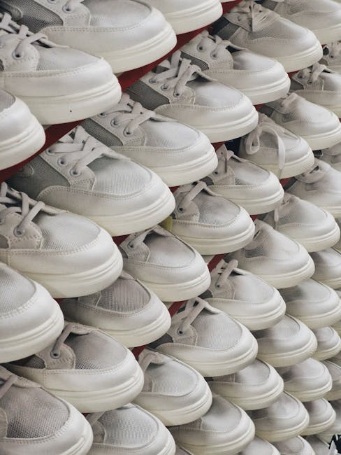 A neatly arranged display of multiple white sneakers, predominantly made of leather and mesh, with rounded toes, white laces, and textured soles, stacked in multiple tiers on a red shelving unit. The shoes show minimal signs of wear and are positioned side by side, filling the frame from the foreground to the background. This arrangement suggests a retail environment or storage area, possibly associated with shoe disposal or clearance, illustrating the management of footwear waste or surplus in a professional setting. The lighting highlights the clean, smooth surfaces of the shoes, contributing to a clear, detailed visual representation. The scene aligns with waste management practices that include sorting, storing, or preparing footwear for disposal, redistribution, or recycling, relevant to alternative waste handling options for footwear waste removal by services like Waste Collection Hackney.