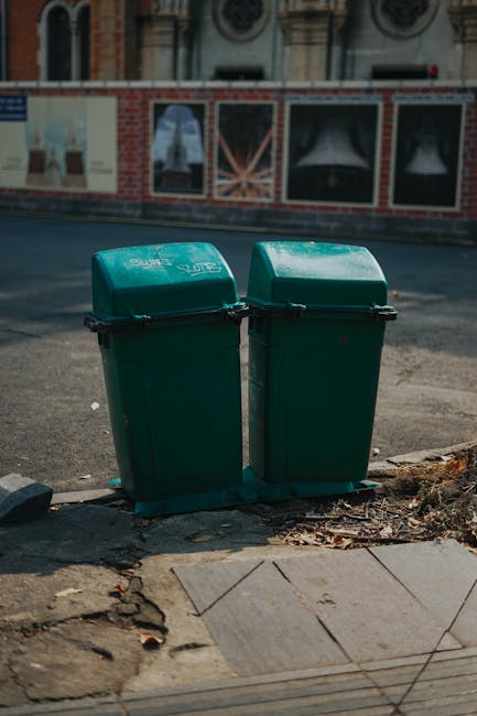 Two green plastic waste bins with hinged lids, positioned side by side on a concrete sidewalk curb edge, with one slightly in front of the other. The bins have a smooth, matte surface with visible black security clips securing the lids. They are situated outdoors on a paved area adjacent to a road, with a rough, uneven edge of the sidewalk visible at the bottom of the image. In the background, there is a street scene featuring a building with ornate windows and a decorative brick wall, along with a painted advertisement on wooden fencing, suggesting an urban environment. The lighting appears natural, with shadows cast across the pavement, indicating daytime. The scene is typical of private waste collection setups often seen in residential or commercial areas, where independent rubbish bins are positioned for curbside collection. The context aligns with waste management practices that involve on-site disposal and separate collection points, as managed by waste collection services like Waste Collection Hackney.